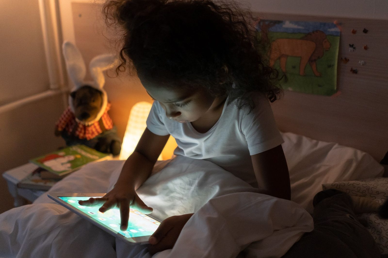 Young child alone in bed, face lit by a tablet screen, in their bedroom at night