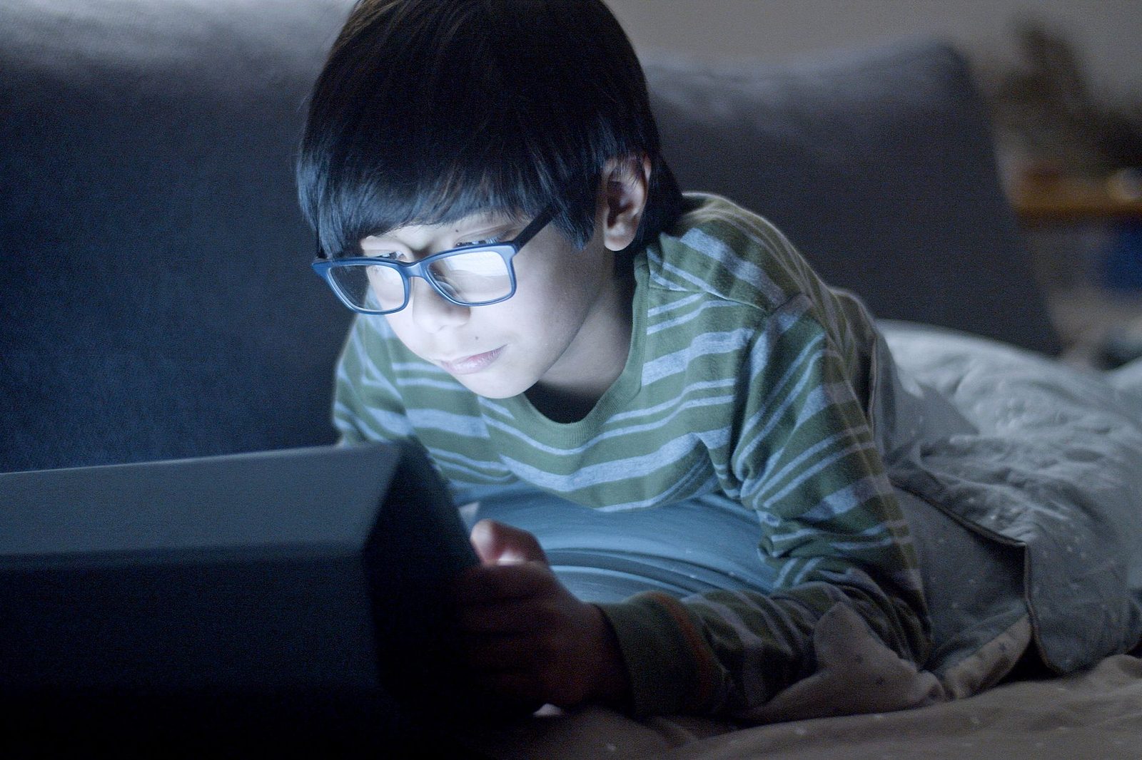 Young boy with glasses lying on a bed, face lit by a tablet screen in the dark