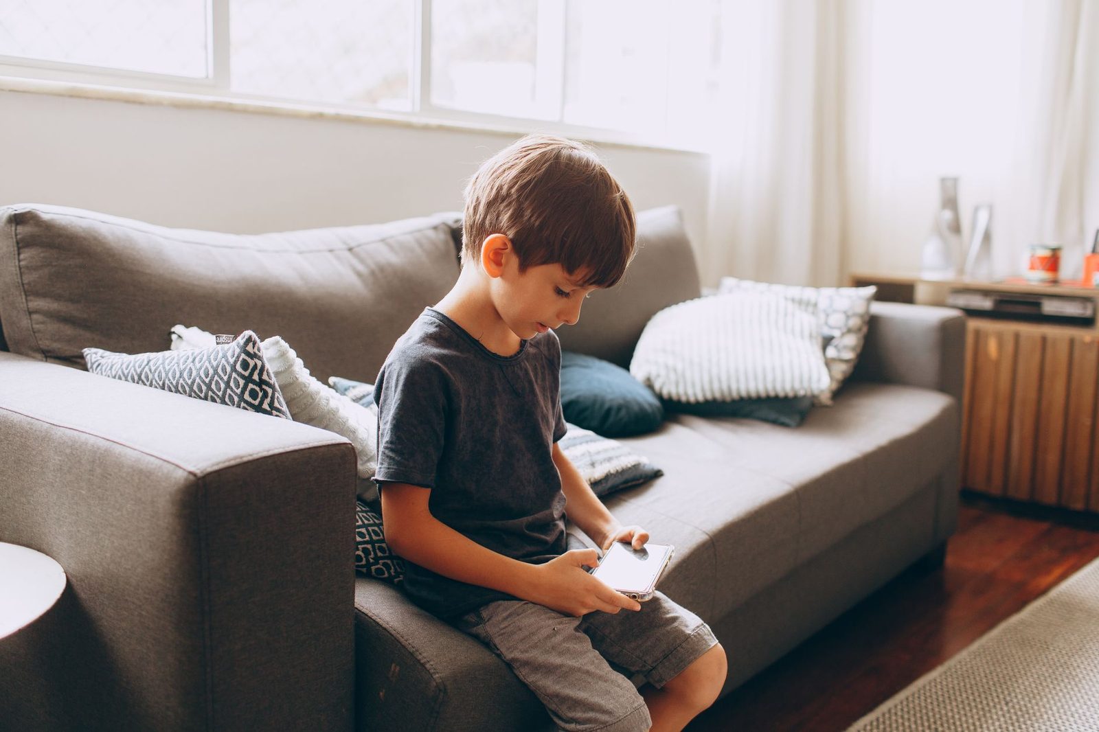 Young boy sitting alone on a sofa in a living room, looking at a smartphone screen