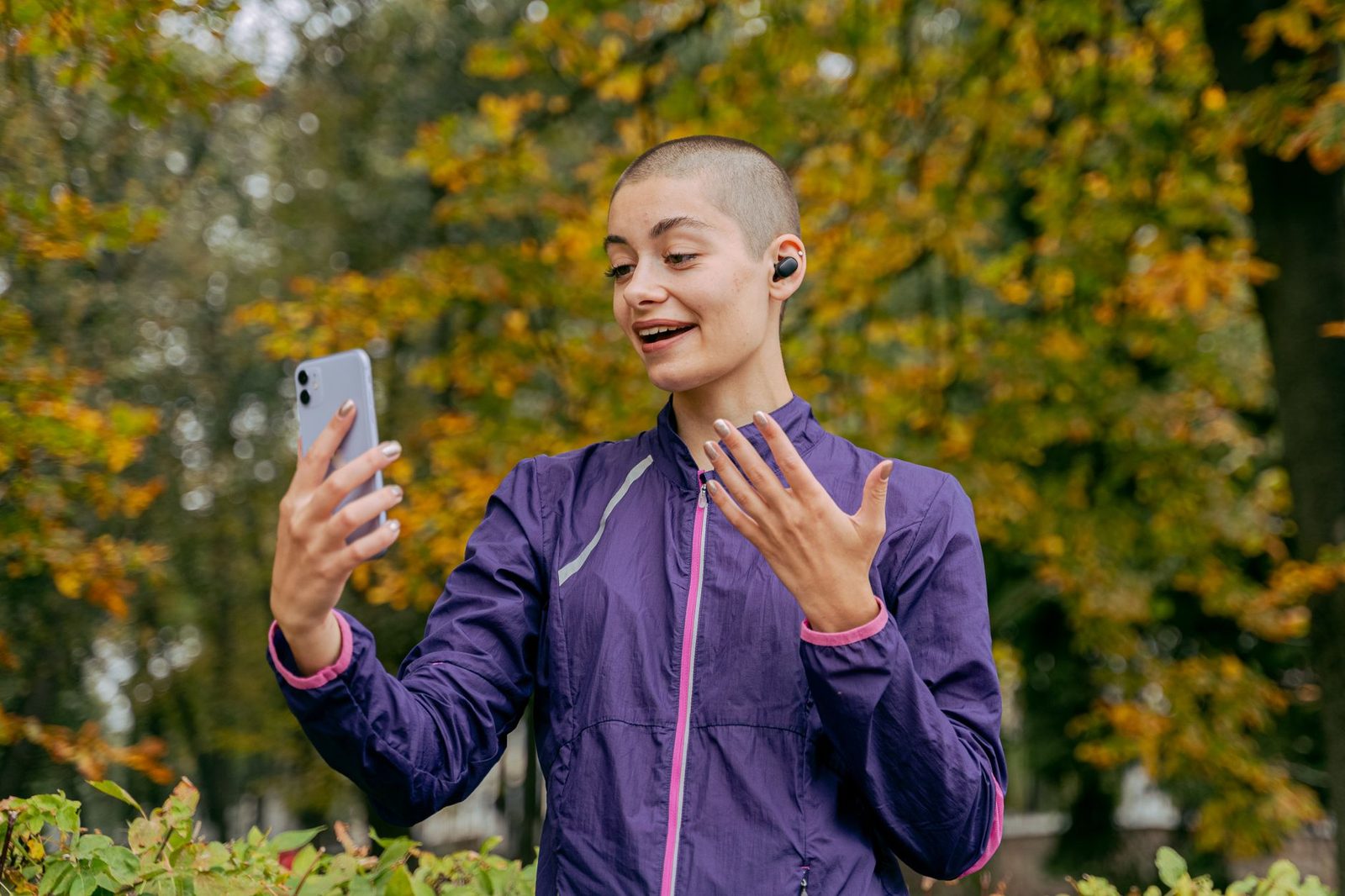 Jeune femme en veste violette souriant lors d'un appel vidéo sur son smartphone dans un parc en automne