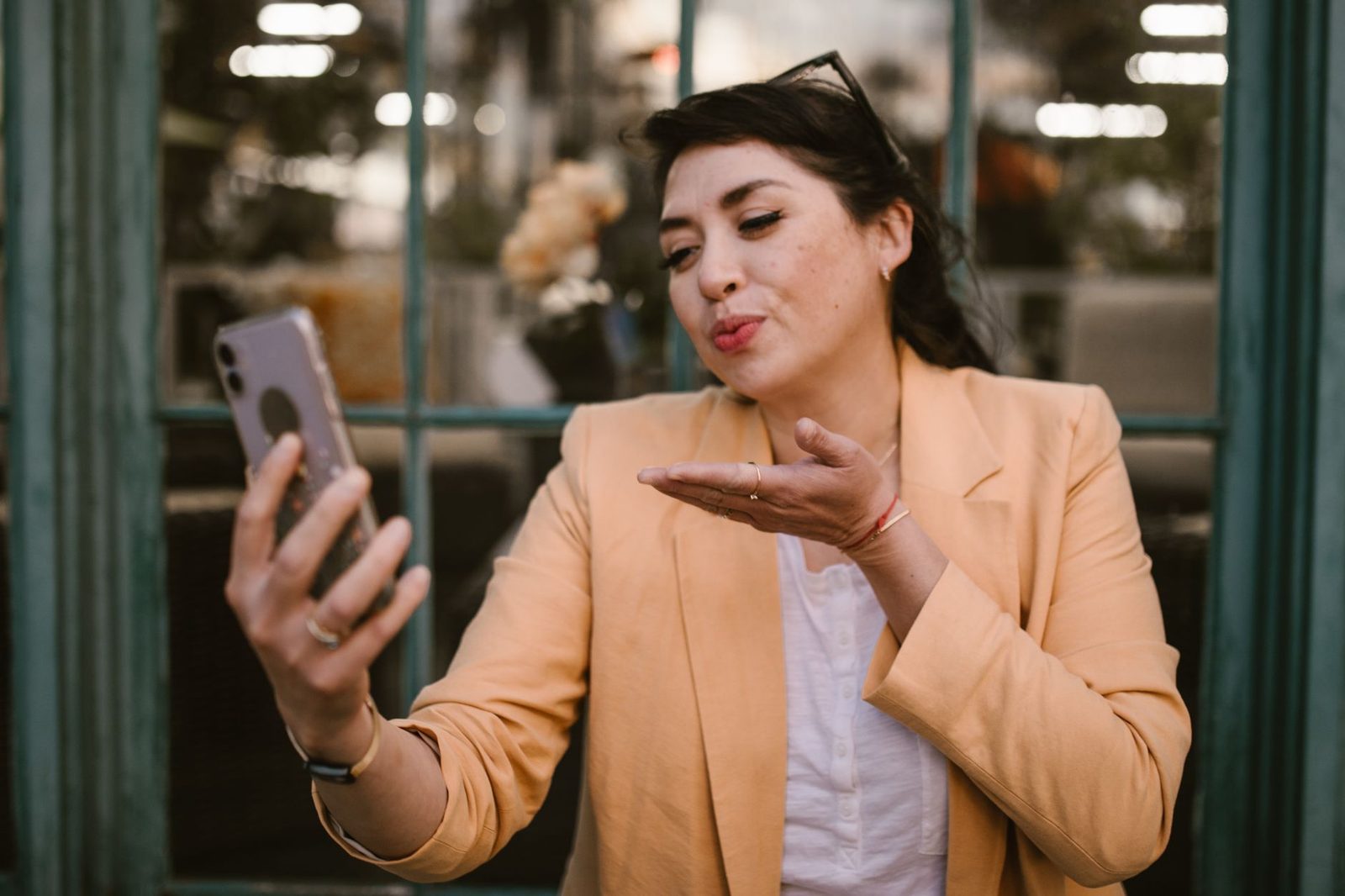 Femme en veste beige envoyant un baiser lors d'un appel vidéo sur son smartphone en extérieur