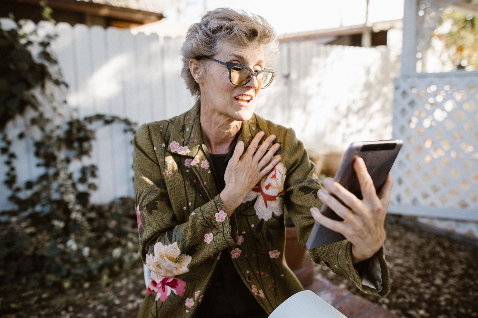 Mujer mayor con gafas y chaqueta de flores reaccionando con emoción durante una videollamada en su tablet al aire libre