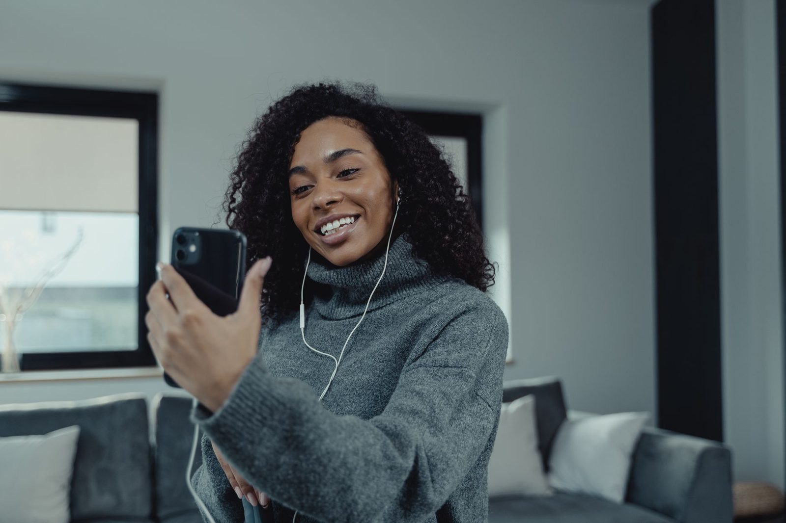 Joven sonriente con auriculares con cable en una videollamada desde su smartphone en un salón
