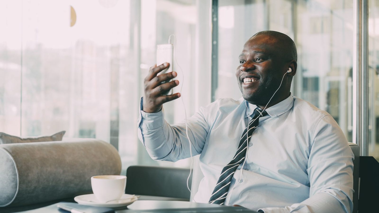 Homme souriant en chemise et cravate passant un appel vidéo sur son smartphone avec des écouteurs, dans un café
