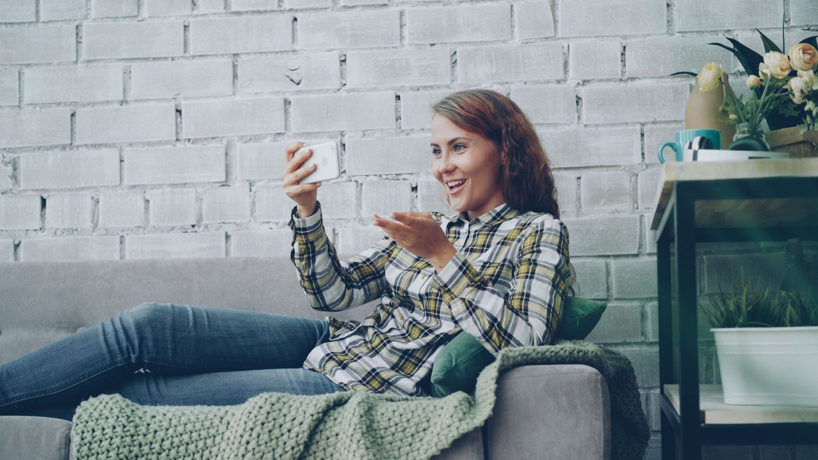 Jeune femme souriante assise sur un canapé, faisant un appel vidéo avec son smartphone dans un salon