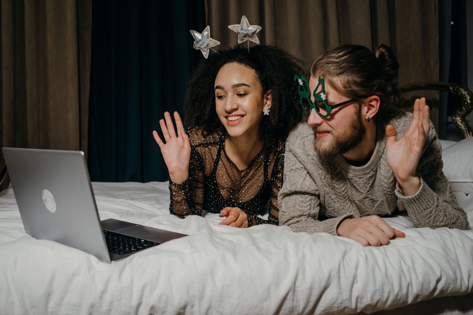 Un couple allongé sur un lit fait signe de la main devant un ordinateur portable lors d'un appel vidéo
