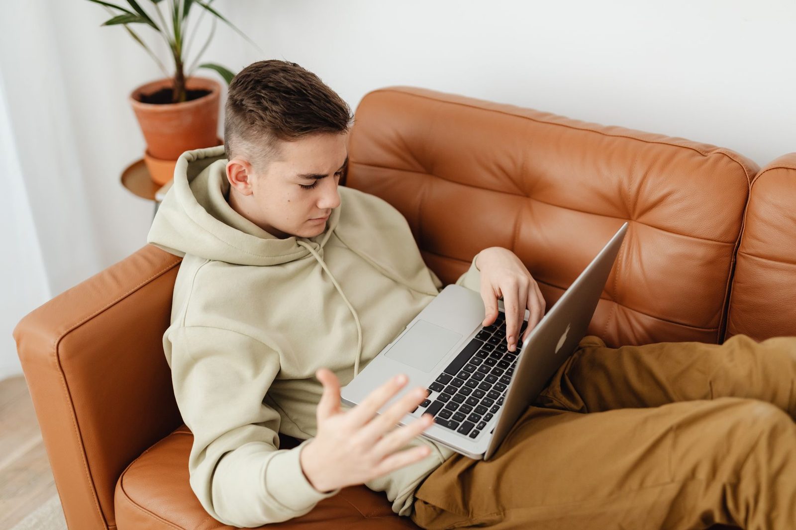 Ragazzo con felpa con cappuccio seduto su un divano in pelle, mentre usa un computer portatile