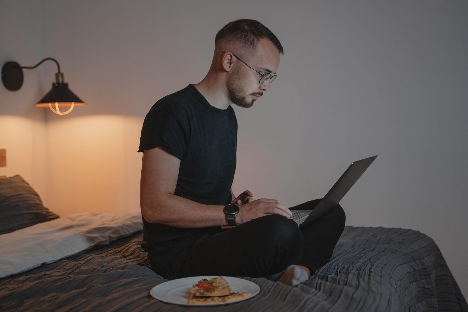 Mann im Schneidersitz auf einem Bett am Abend, mit einem Laptop neben einem Teller Pizza
