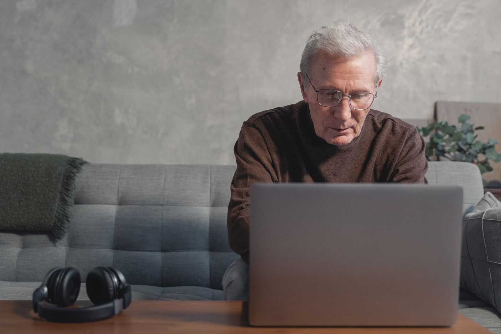 Älterer Mann mit Brille auf einem Sofa vor einem Laptop, Kopfhörer auf dem Tisch abgelegt