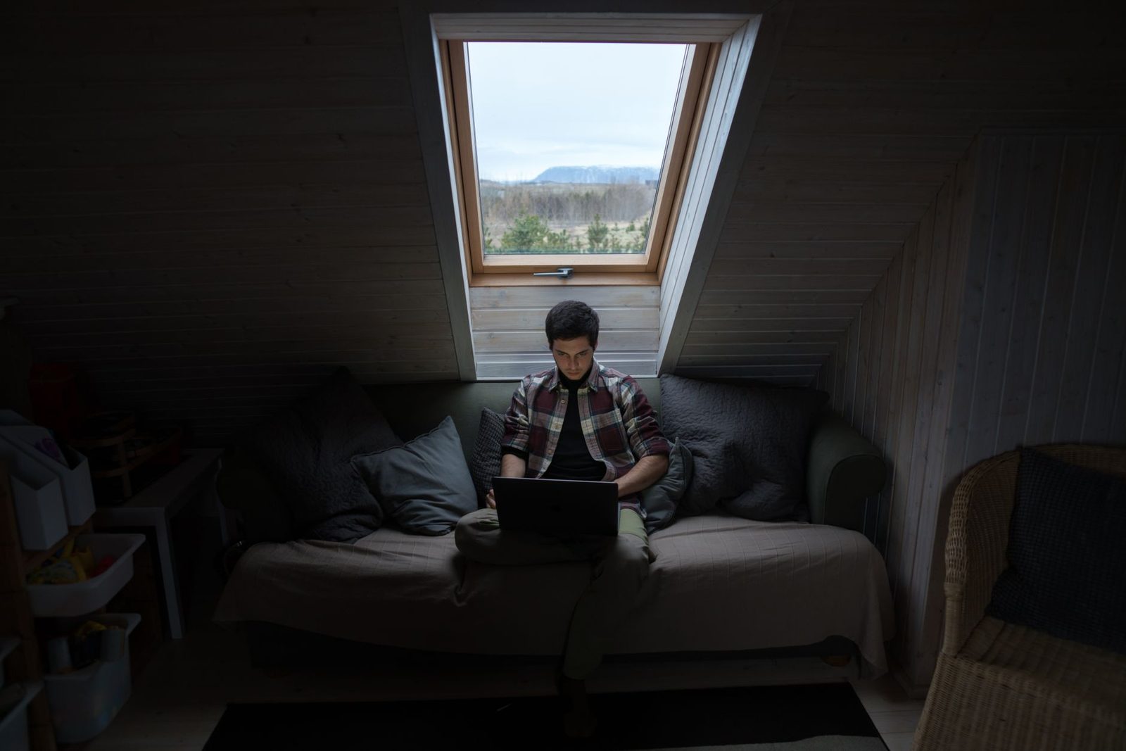 Junger Mann allein auf einem Sofa in einem dunklen Dachzimmer, das Gesicht vom Bildschirm eines Laptops erhellt