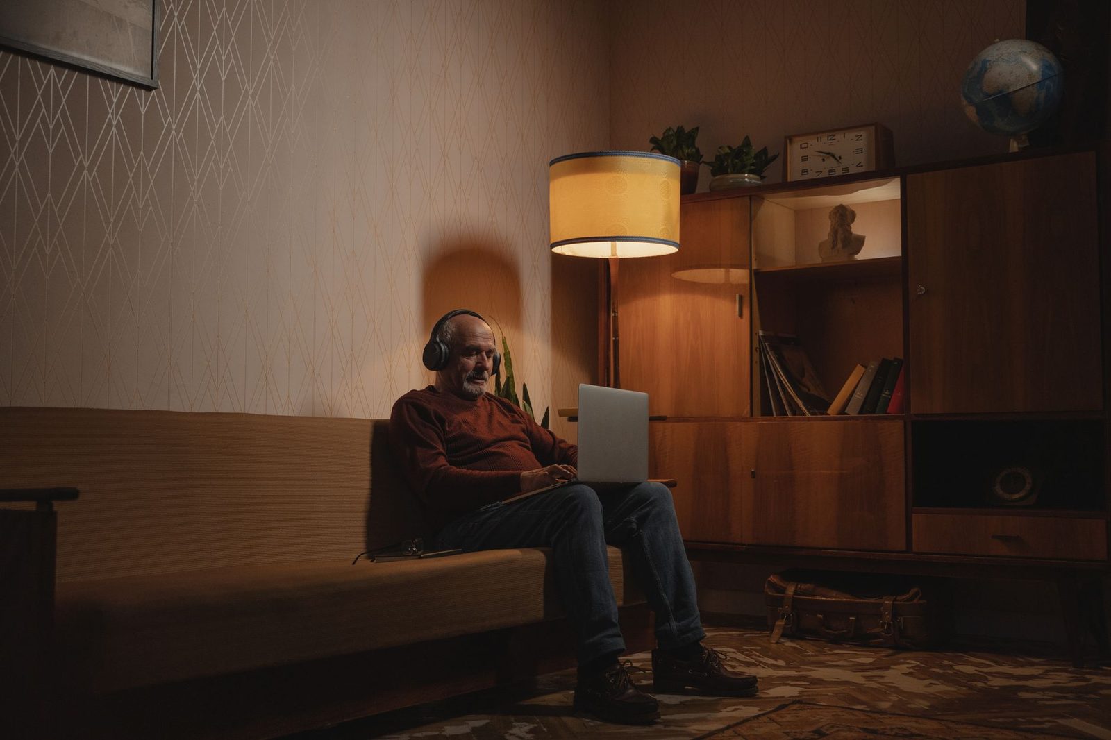 Homme avec casque audio assis sur un canapé, ordinateur portable sur les genoux, dans un salon éclairé par une lampe