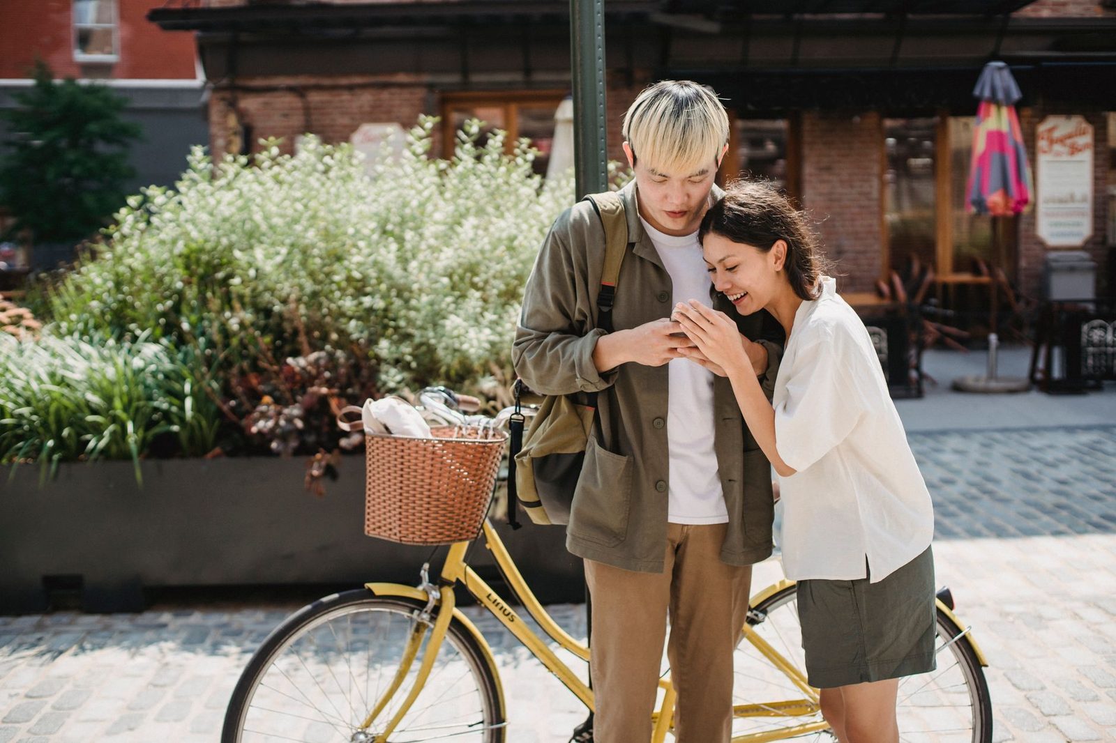 Un jeune couple souriant regarde ensemble un smartphone dans une rue pavée, près d'un vélo jaune