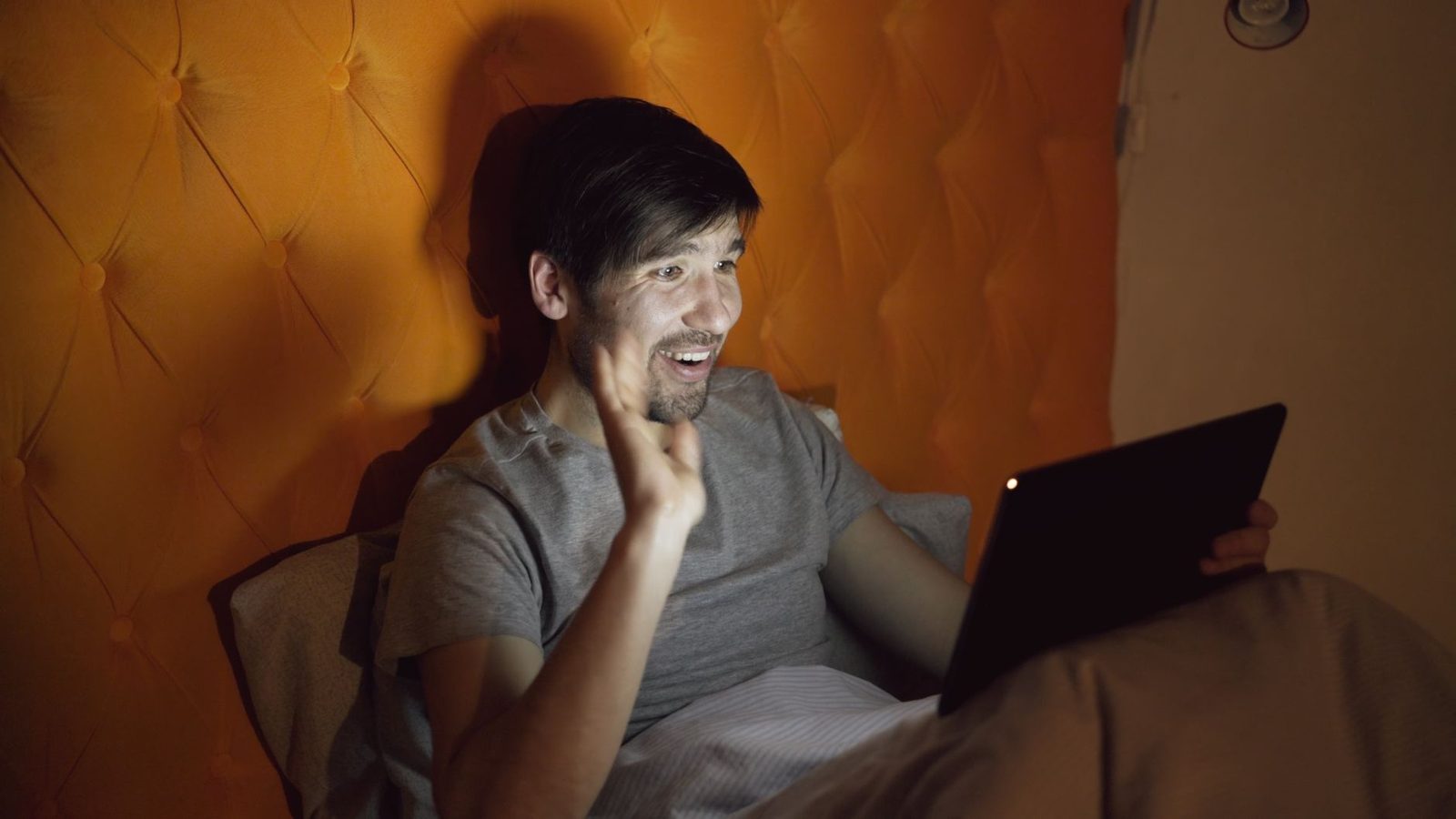 Homme souriant faisant un signe de la main devant une tablette, allongé dans son lit le soir
