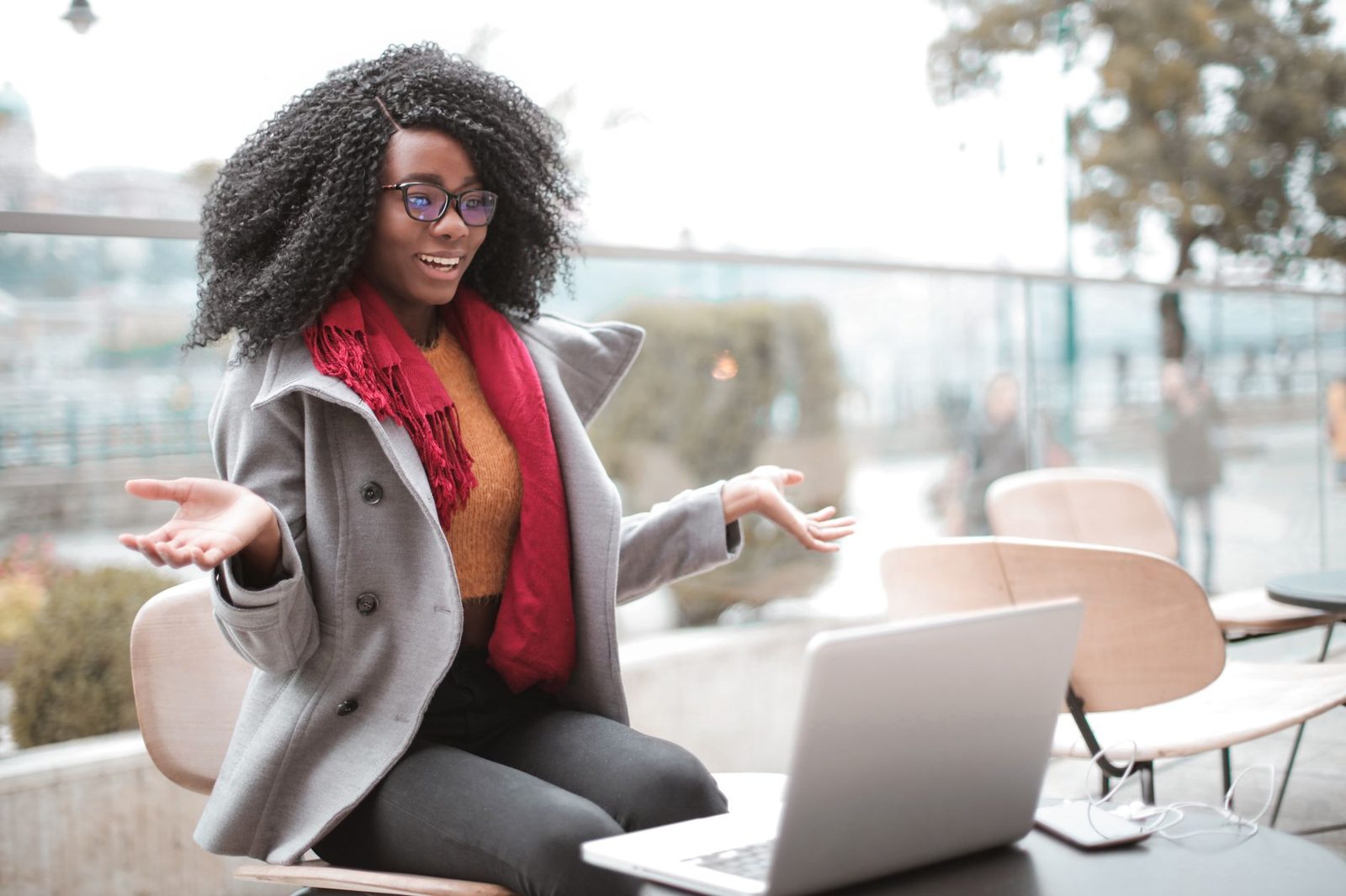Jeune femme souriante à lunettes, en terrasse, gesticulant devant un ordinateur portable
