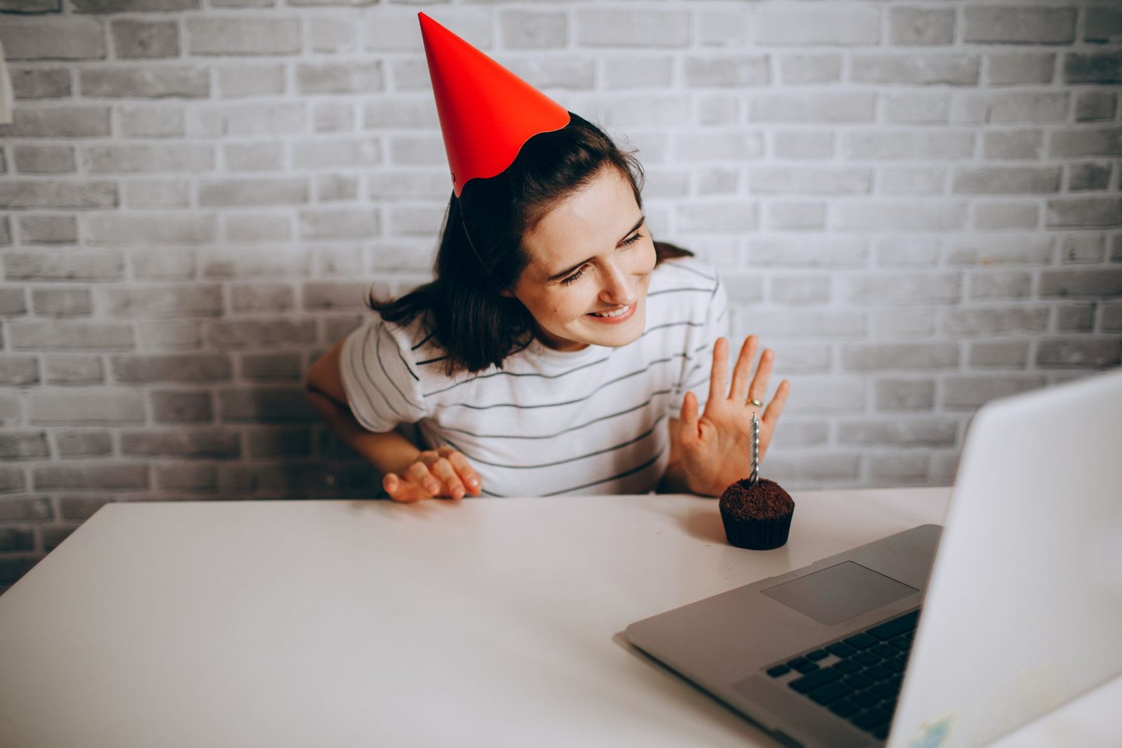 Femme avec un chapeau de fête et un cupcake saluant de la main devant l'écran d'un ordinateur portable