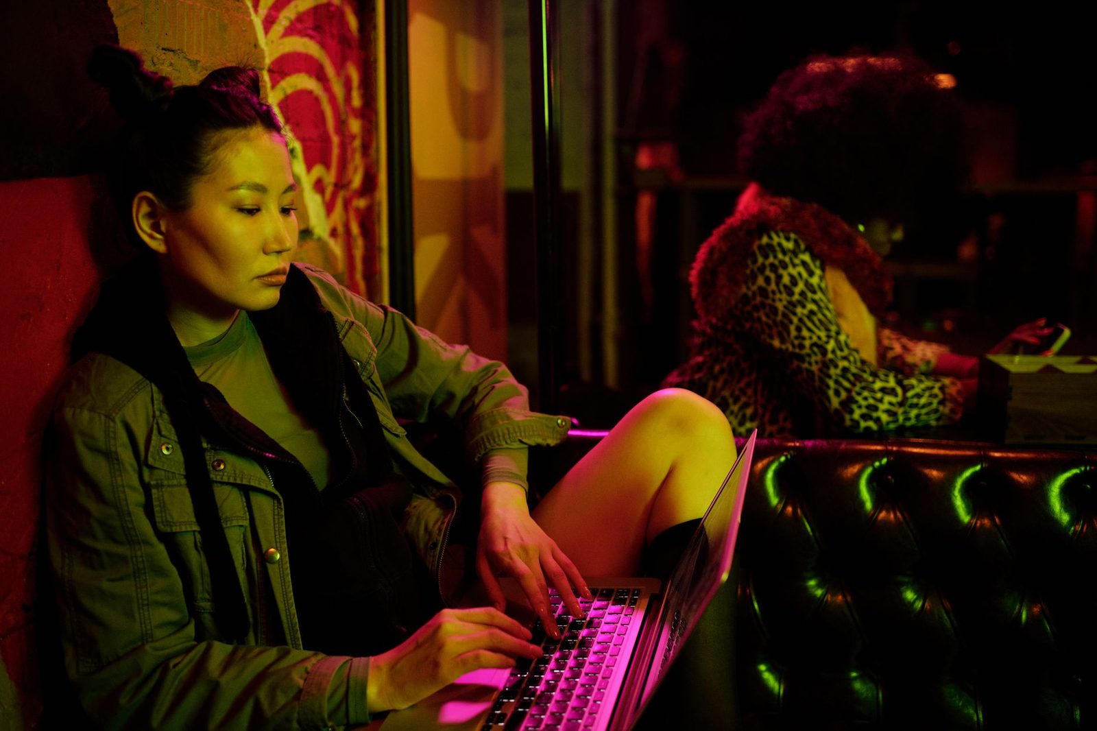 Young woman sitting and typing on a laptop in a dark room lit by colourful neon lights