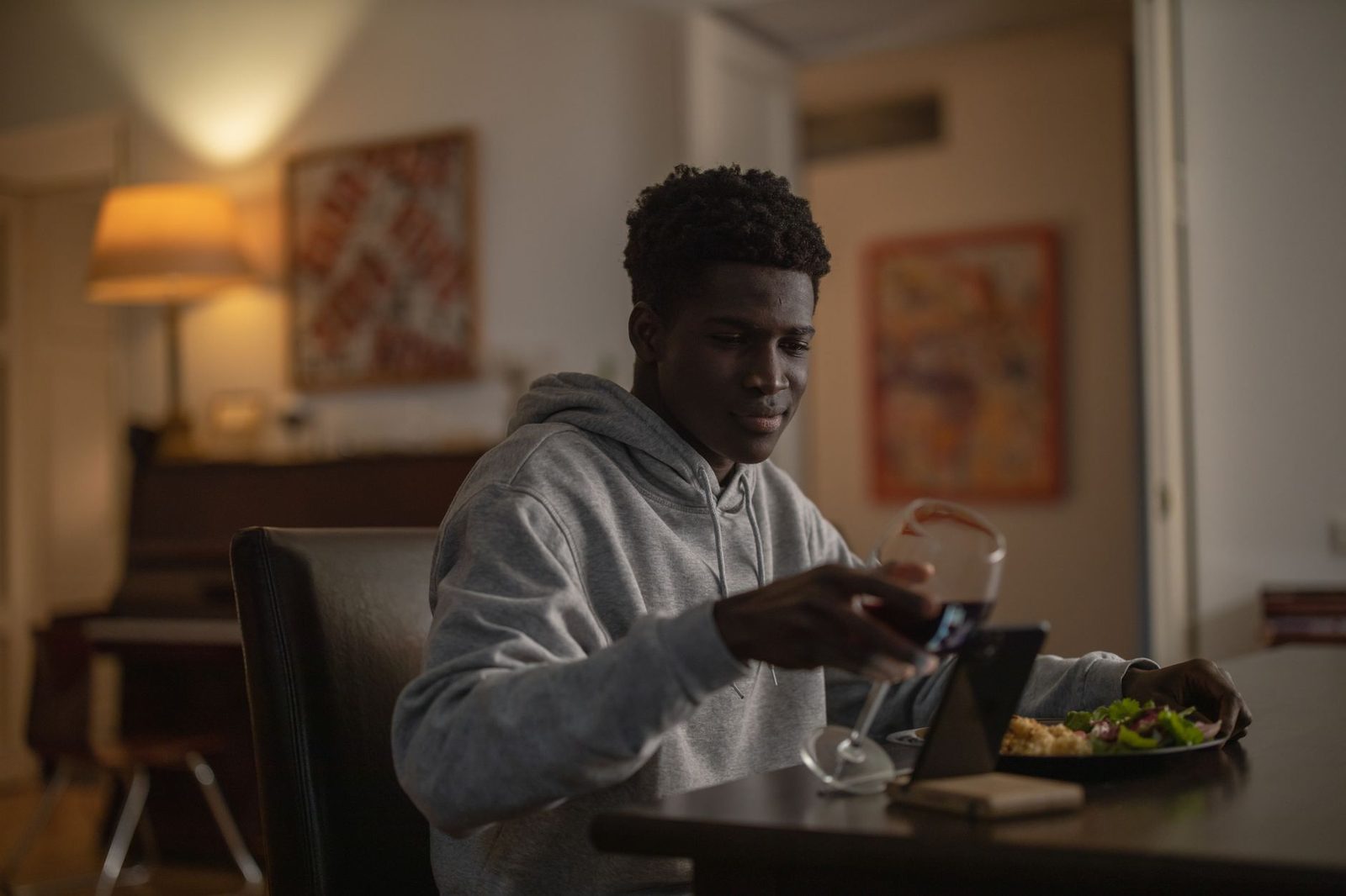 Jeune homme en sweat gris tenant un verre de vin devant une tablette lors d'un appel vidéo à table
