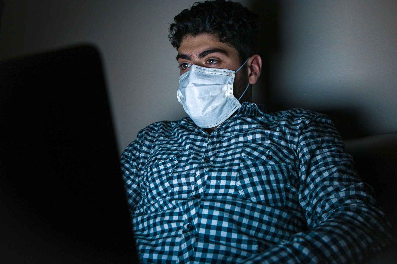 Homme portant un masque chirurgical devant un ordinateur portable dans une pièce sombre