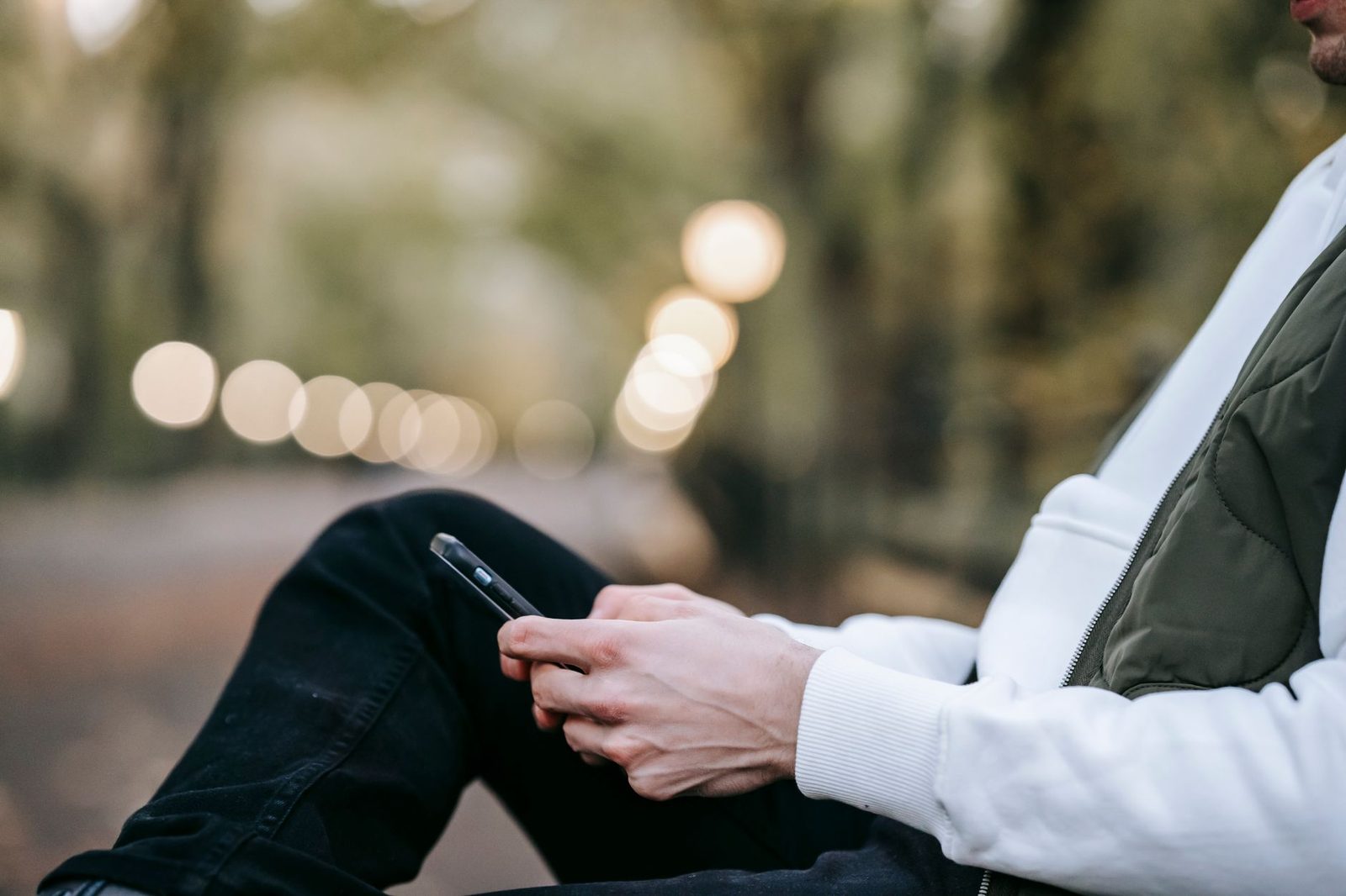Homem sentado no exterior, de colete, a usar um smartphone, com luzes desfocadas ao fundo