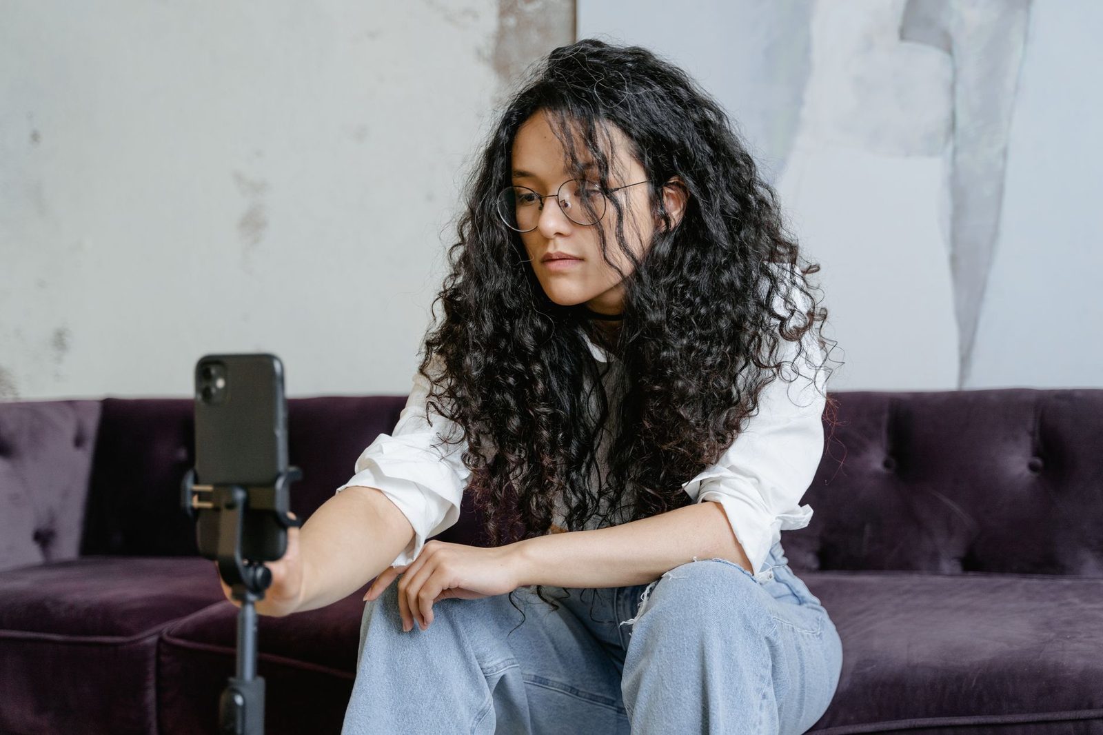Jovem de cabelo encaracolado sentada num sofá, a filmar com um smartphone num tripé