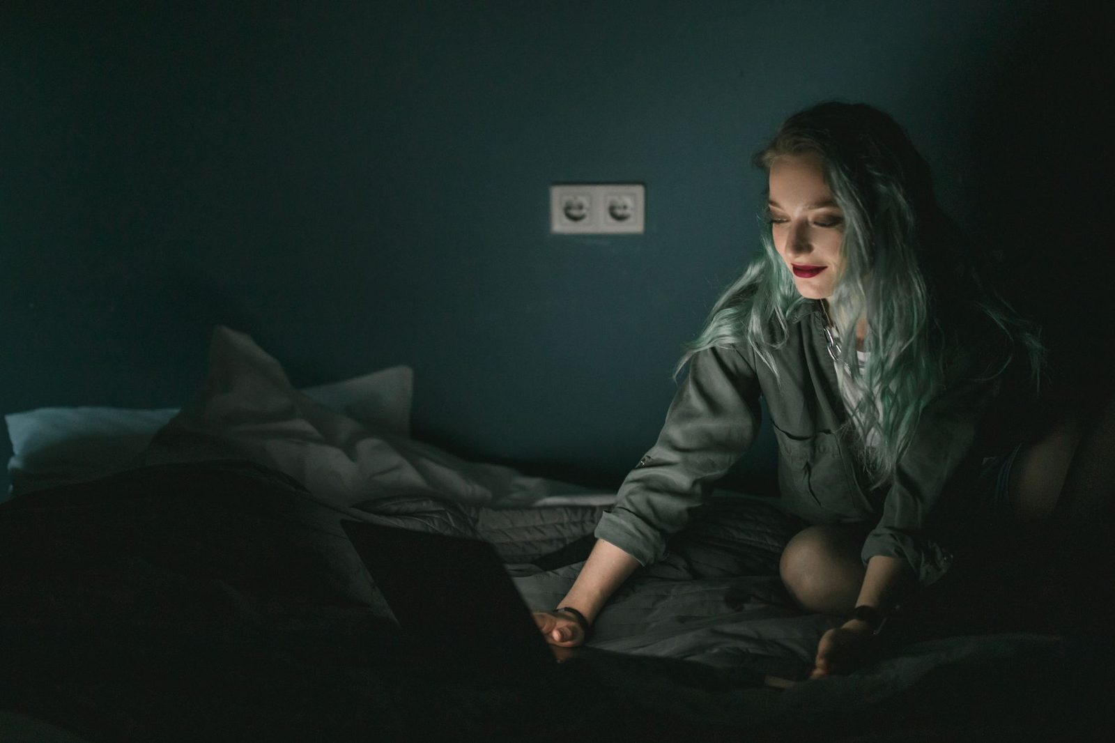 Jeune femme aux cheveux clairs assise sur un lit dans une chambre sombre, devant un ordinateur portable allumé