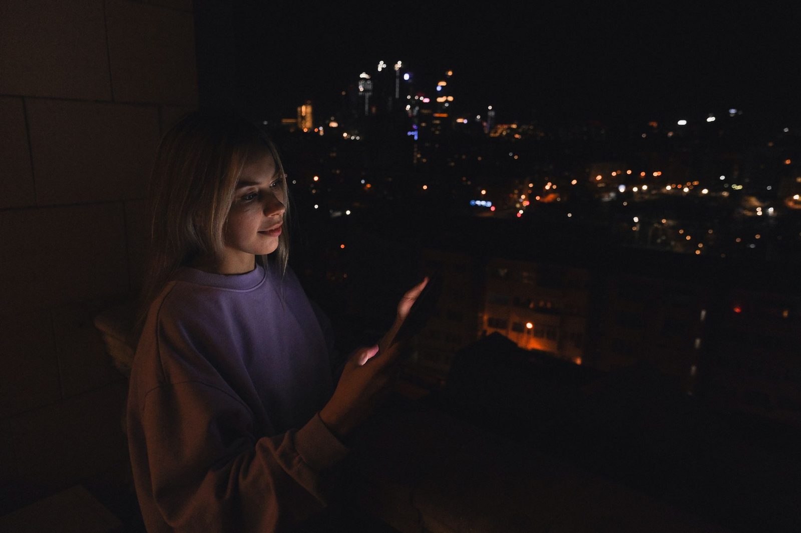 Jeune femme utilisant son smartphone la nuit sur un balcon, lumières de la ville en arrière-plan