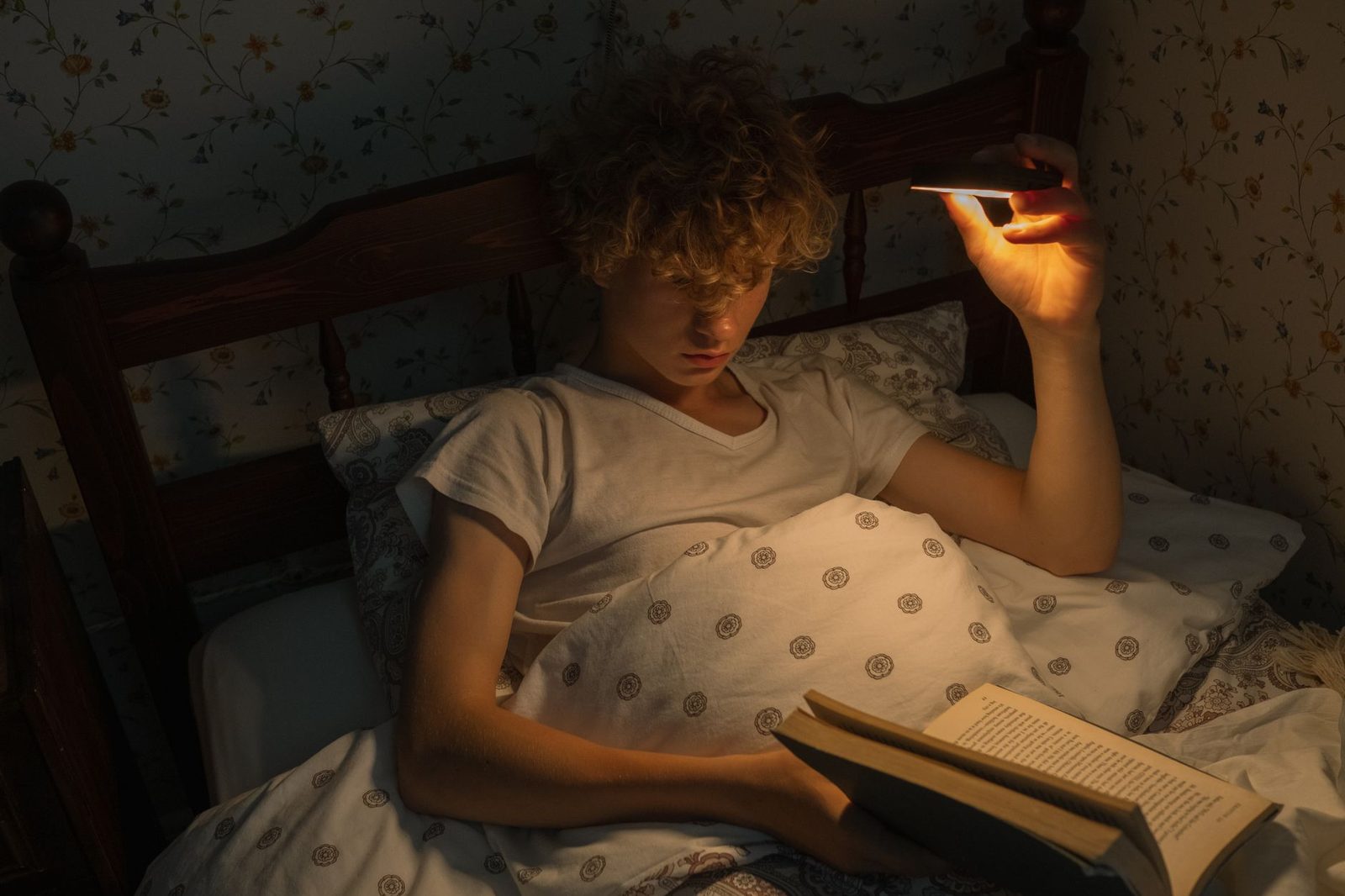 Jugendlicher nachts im Bett, der ein eingeschaltetes Smartphone über ein aufgeschlagenes Buch hält