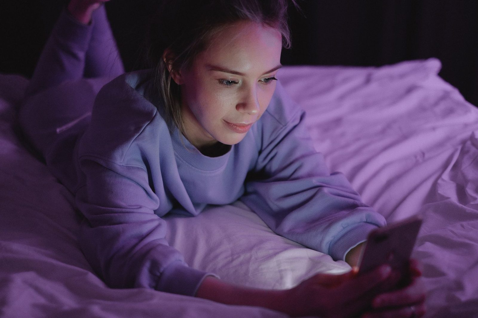 Junge Frau, die im Halbdunkel auf einem Bett liegt, ihr Gesicht vom Bildschirm ihres Smartphones erhellt