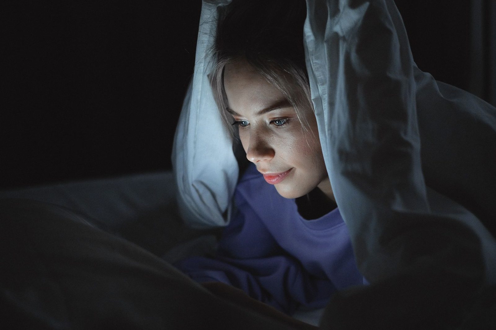Jeune femme allongée sous la couette dans le noir, le visage éclairé par l'écran de son téléphone