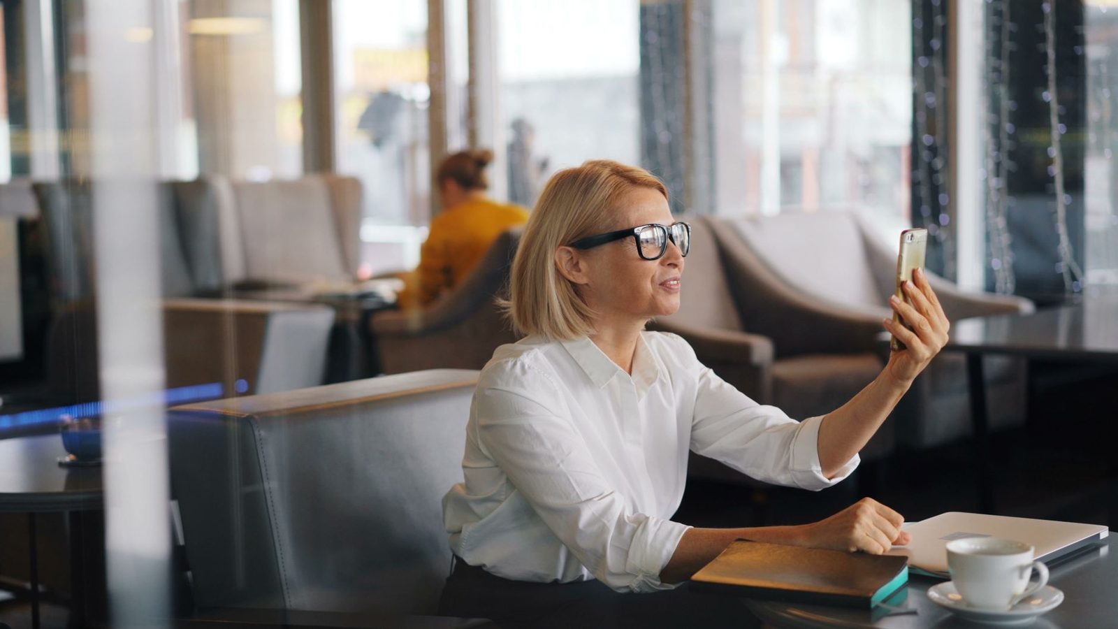 Femme blonde à lunettes en chemise blanche lors d'un appel vidéo sur smartphone dans un café