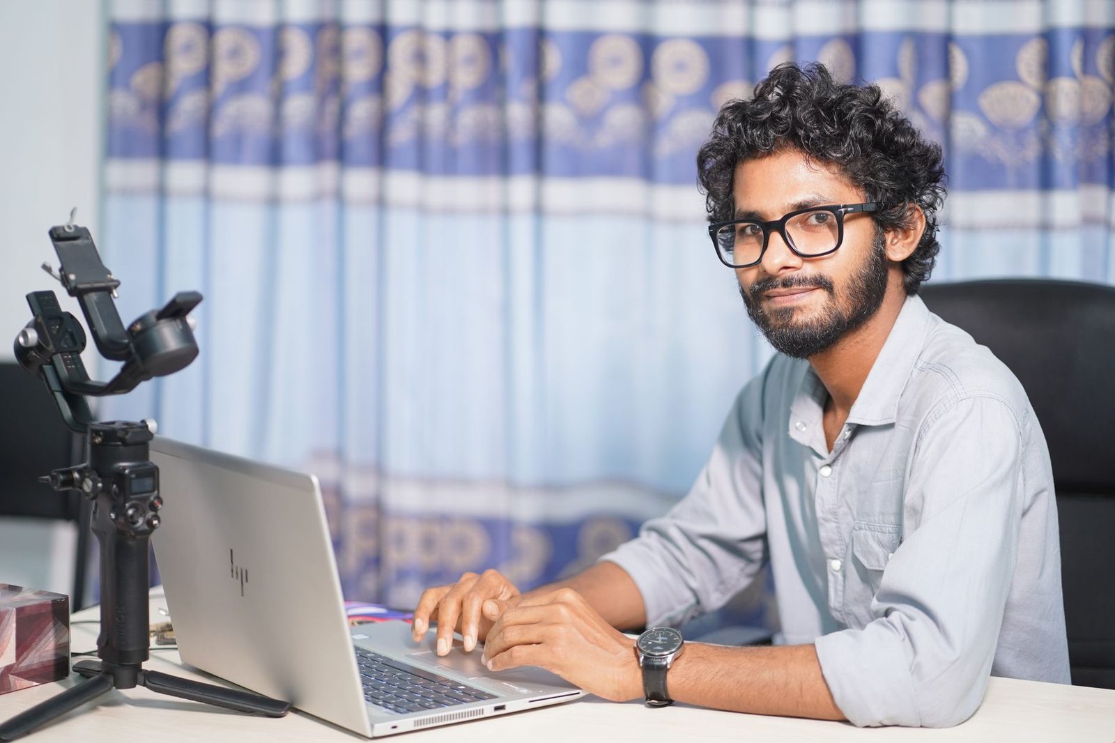 Junger Mann mit Brille sitzt vor einem Laptop und einer Kamera und lächelt in die Linse