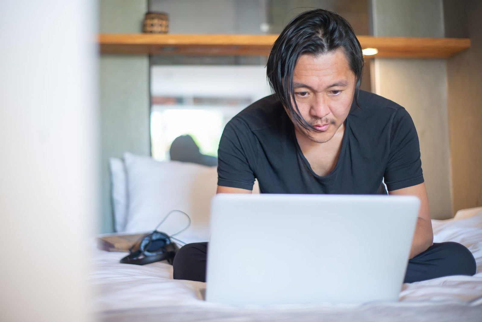 Mann sitzt im Schneidersitz auf einem Bett und schaut auf den Bildschirm seines weißen Laptops