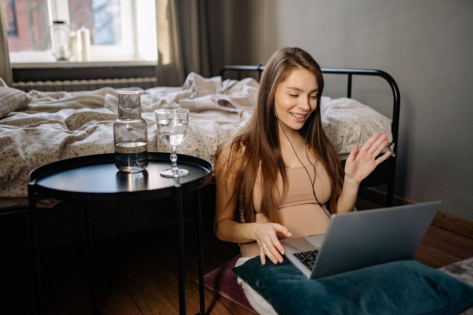 Jeune femme souriante avec écouteurs saluant de la main devant un ordinateur portable dans sa chambre