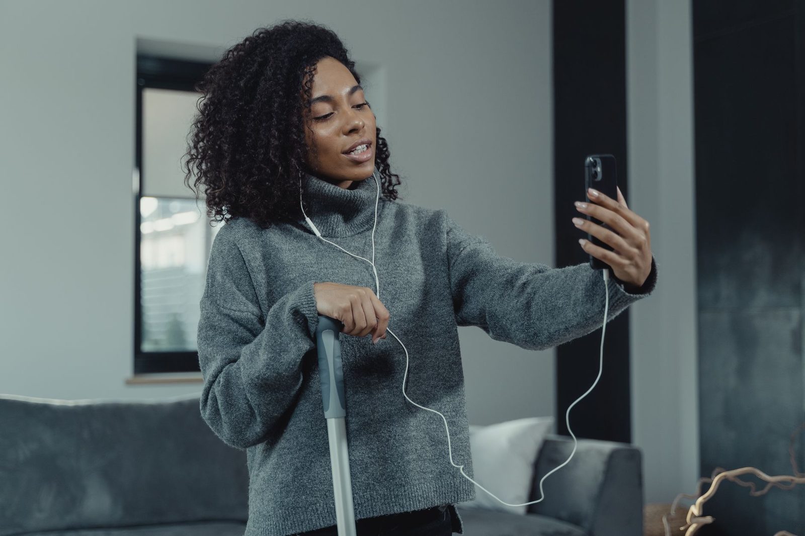 Jeune femme en pull gris avec écouteurs filaires en appel vidéo sur son smartphone dans un salon