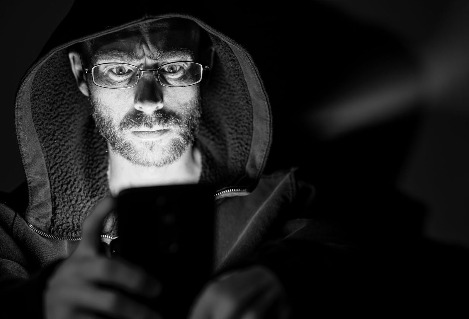 Photo en noir et blanc d'un homme à lunettes, capuche relevée, regardant l'écran de son smartphone dans l'obscurité