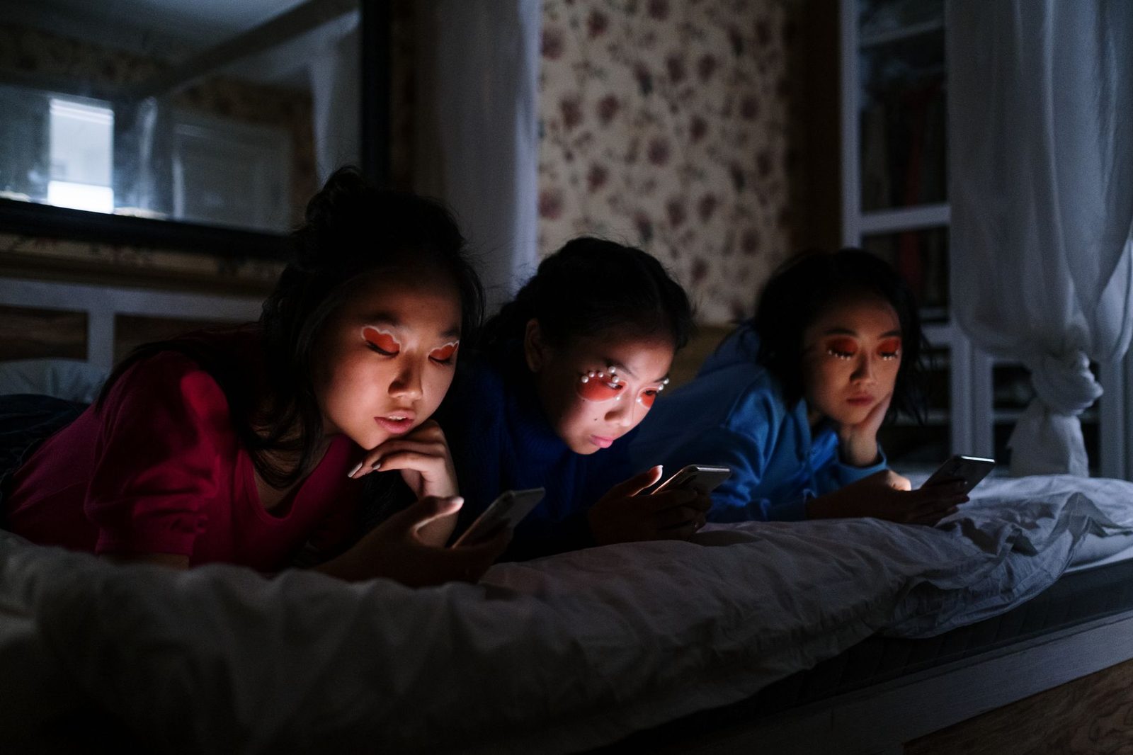 Trois jeunes filles allongées sur un lit dans le noir, regardant l'écran lumineux de leur smartphone