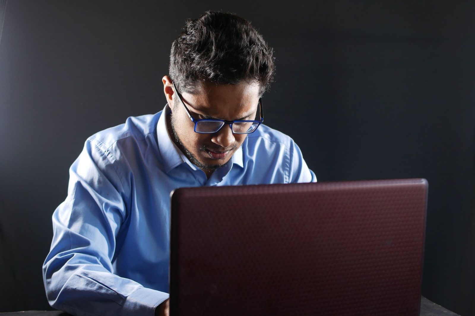 Homme à lunettes en chemise bleue devant un ordinateur portable, sur fond noir