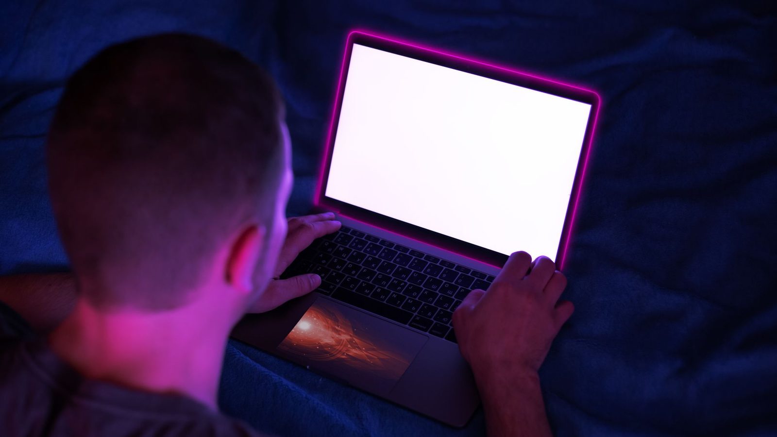 Homme allongé dans le noir devant un ordinateur portable à l'écran blanc, éclairé d'une lueur rose