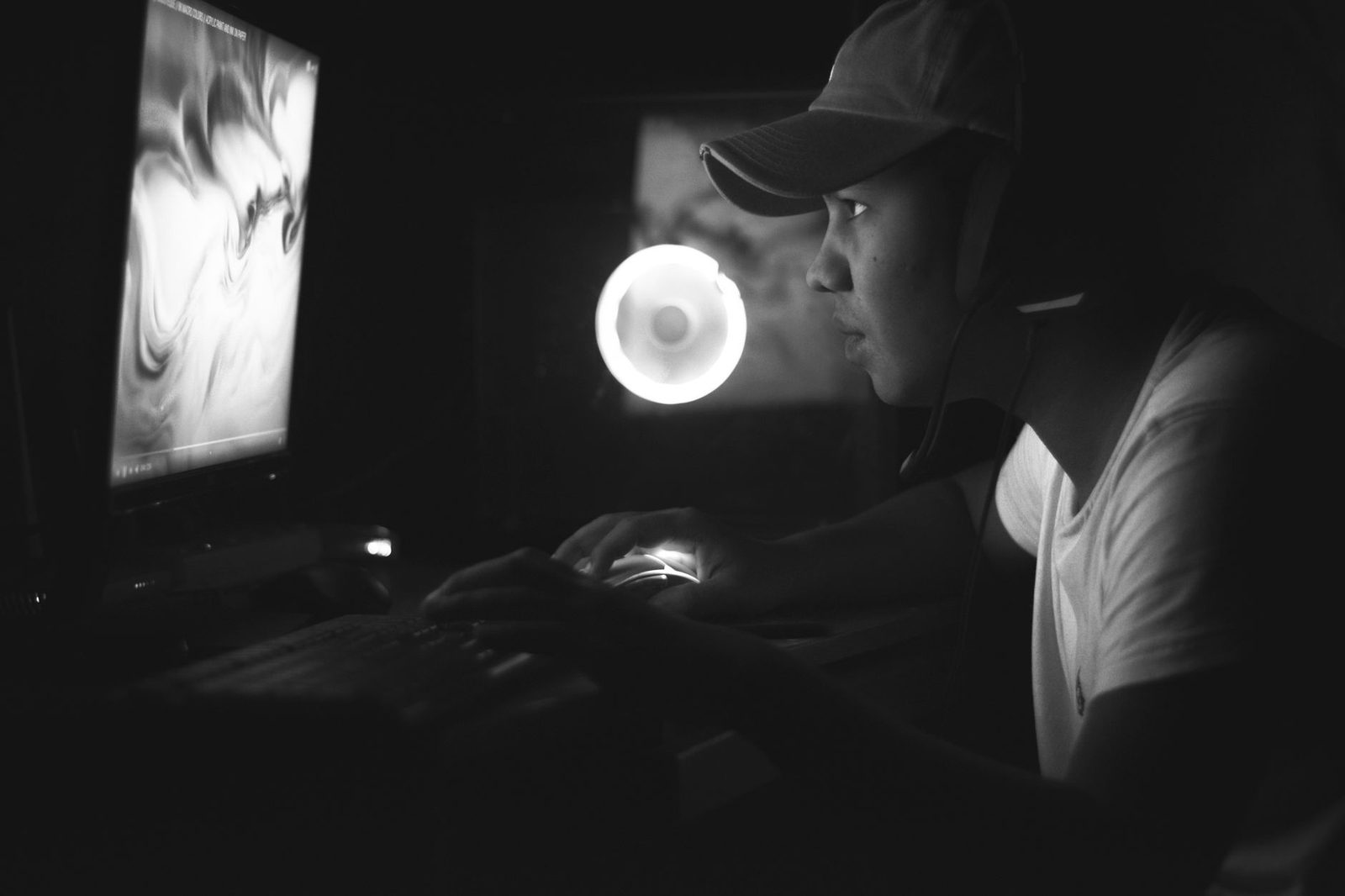 Photo en noir et blanc d'un jeune homme avec casquette et casque devant un écran d'ordinateur dans le noir
