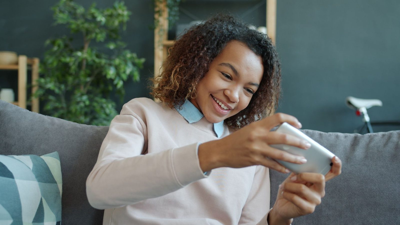Giovane donna sorridente seduta su un divano, che tiene uno smartphone in orizzontale davanti al viso
