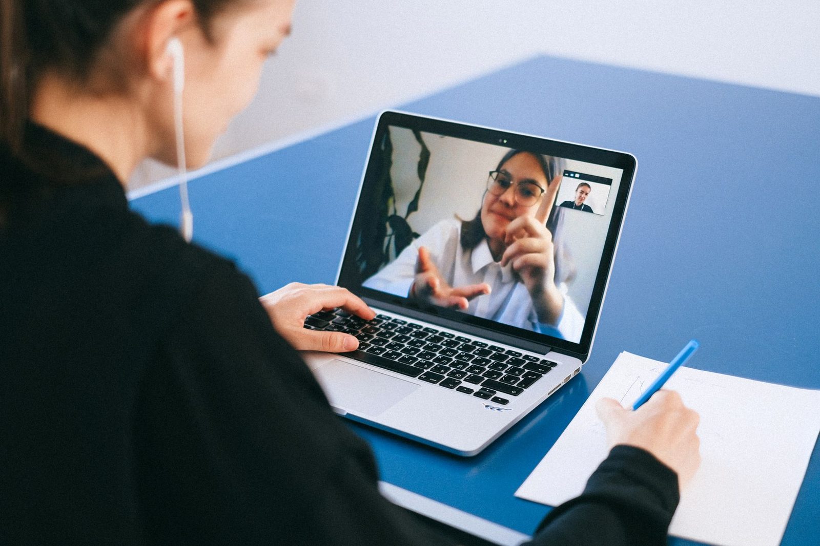 Giovane donna con auricolari in videochiamata su un computer portatile, che prende appunti su una scrivania blu