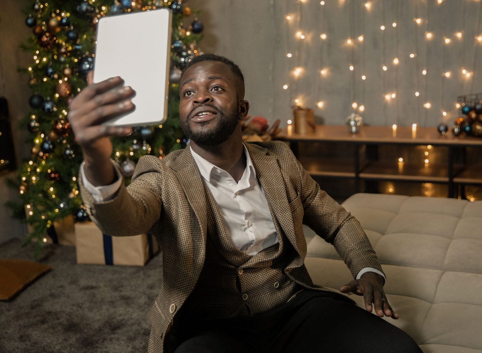 Homme en veste tenant une tablette à bout de bras pour un appel vidéo, devant un sapin et des guirlandes
