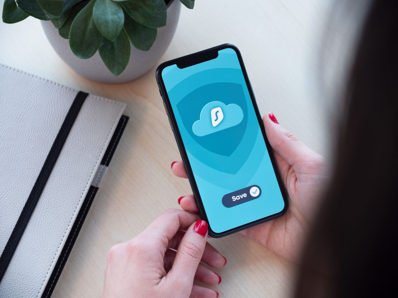Hände halten ein Smartphone mit geöffneter VPN-App auf einem Holzschreibtisch, daneben ein Notizbuch und eine Pflanze