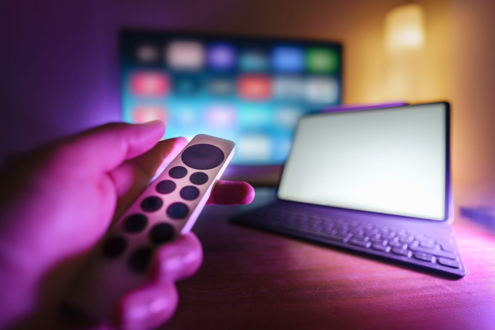 Main tenant une télécommande devant une télévision connectée floue et une tablette avec clavier