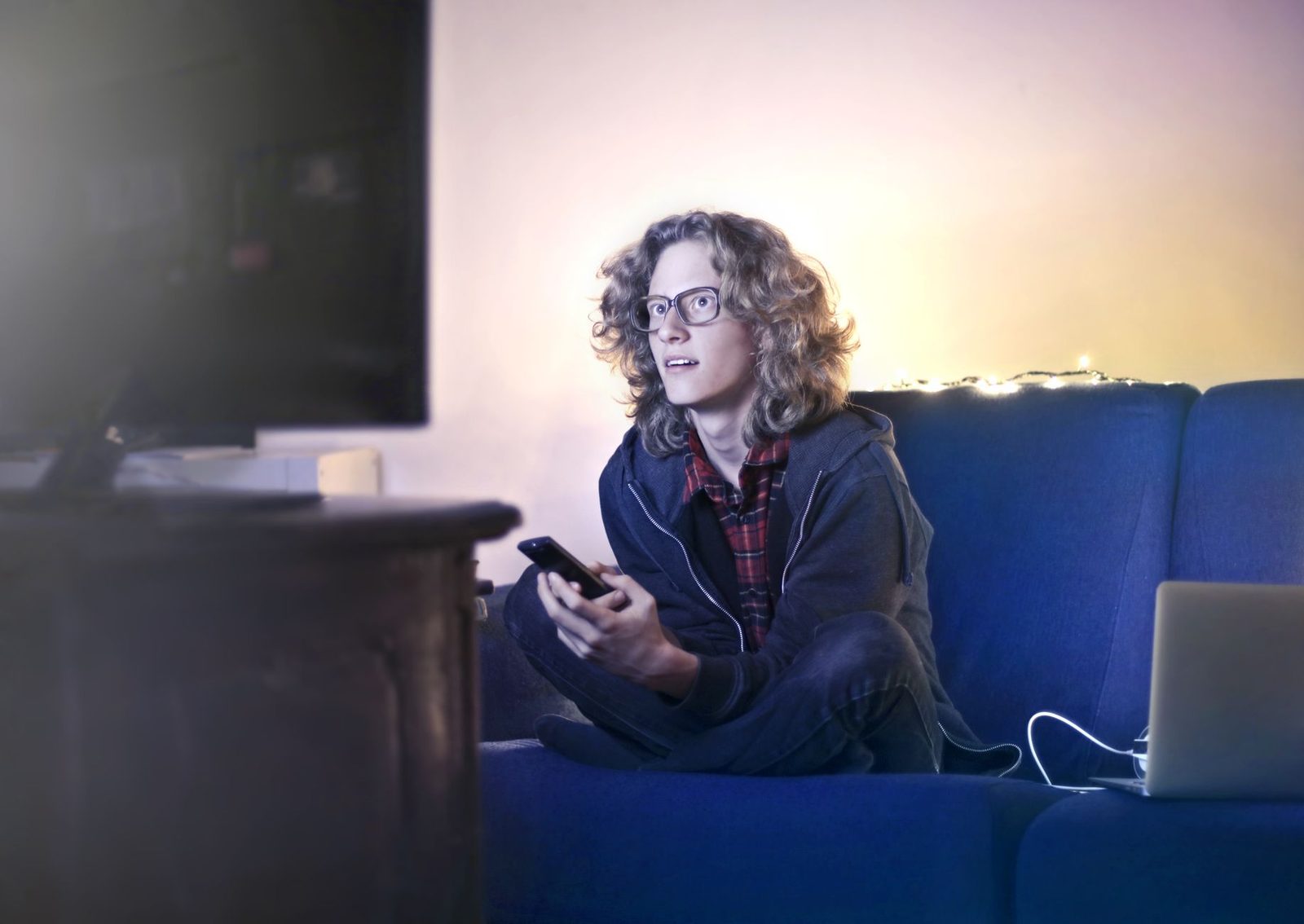 Jeune homme à lunettes assis sur un canapé bleu avec une télécommande devant un écran, un ordinateur portable à côté