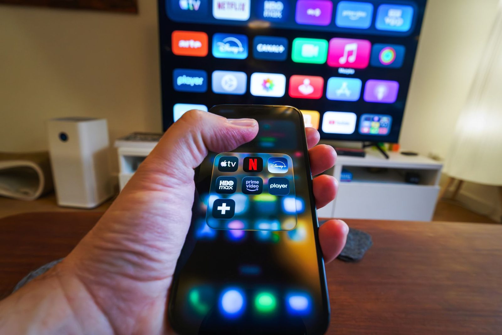 Main tenant un smartphone affichant des applications de streaming devant un téléviseur connecté allumé