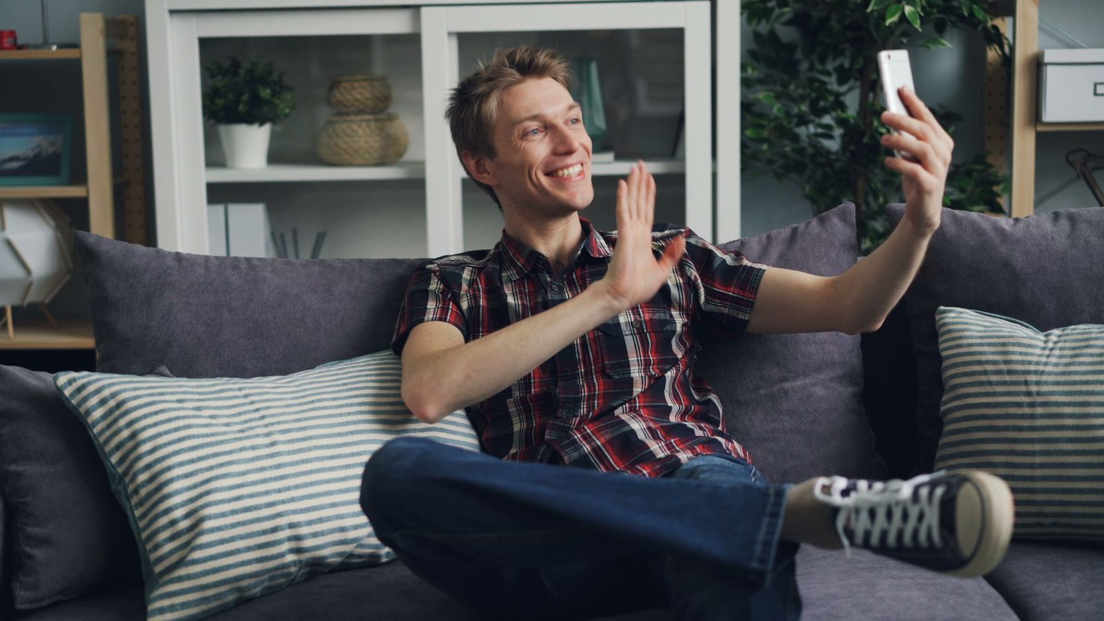 Un homme souriant assis sur un canapé, prenant un selfie ou passant un appel vidéo avec son smartphone.