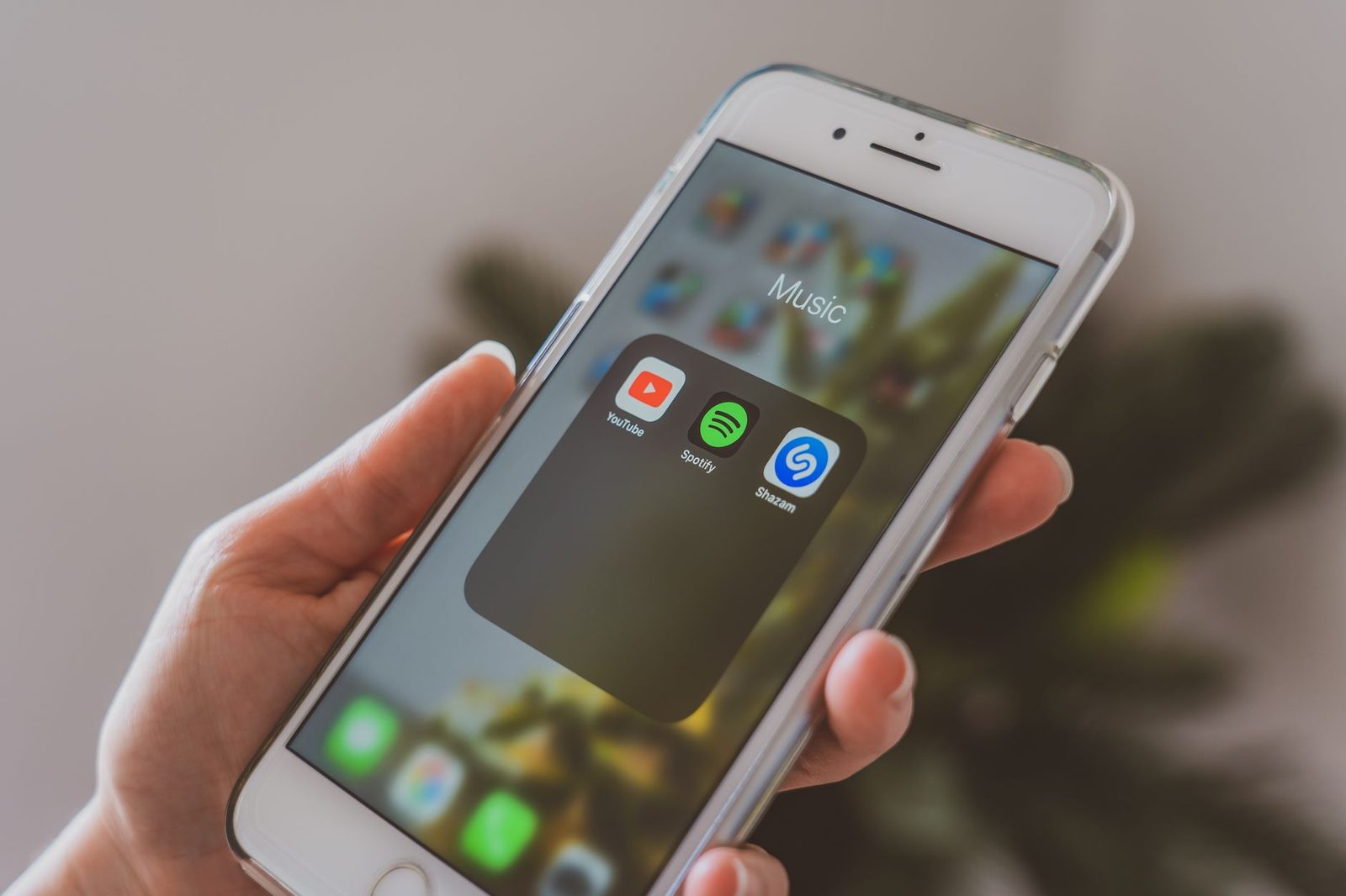 Mano sosteniendo un smartphone que muestra una carpeta de aplicaciones de streaming: YouTube, Spotify y Shazam