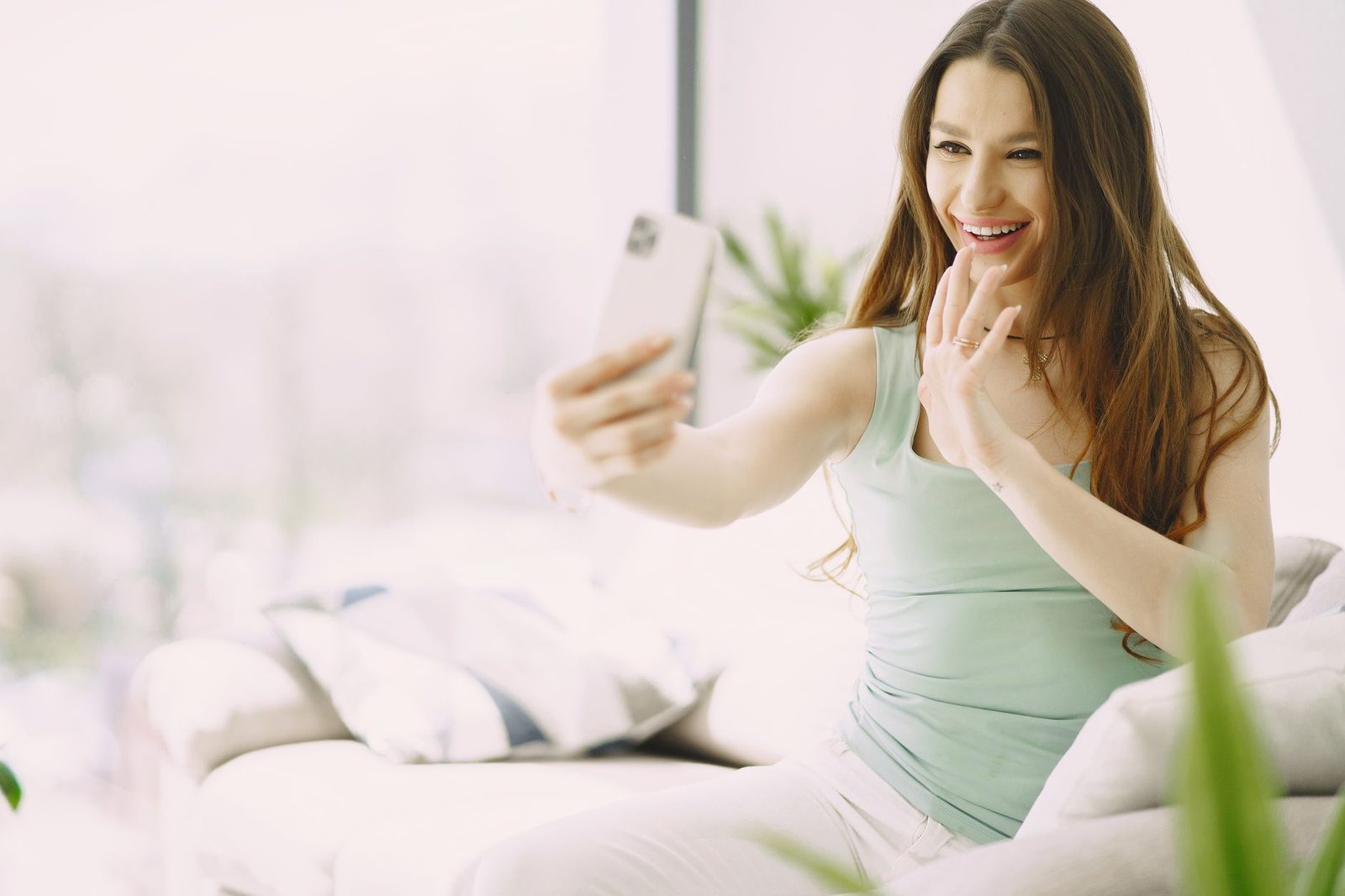 Jeune femme souriante assise sur un canapé, saluant de la main pendant un appel vidéo sur son smartphone